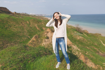 Naklejka premium Young smiling relaxed woman with closed eyes in light casual clothes with headphones listening music clinging to head standing in field near water on green background. Lifestyle, leisure concept.