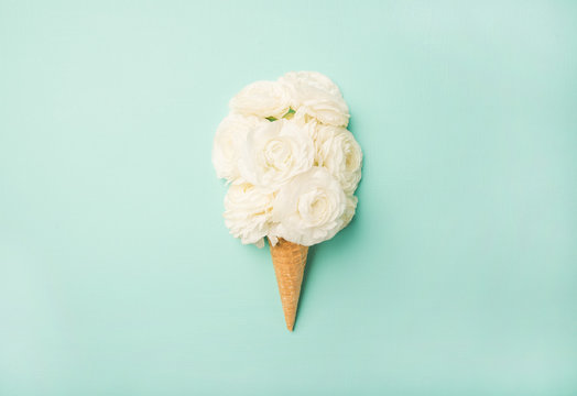 Fototapeta Flat-lay of waffle sweet cone with white buttercup flowers over light blue pastel background, top view. Spring or summer mood concept