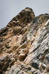 Big mountain cliff under cloudy sky close-up. Beautiful rocky gray textured background with vegetation. Majestic nature.
