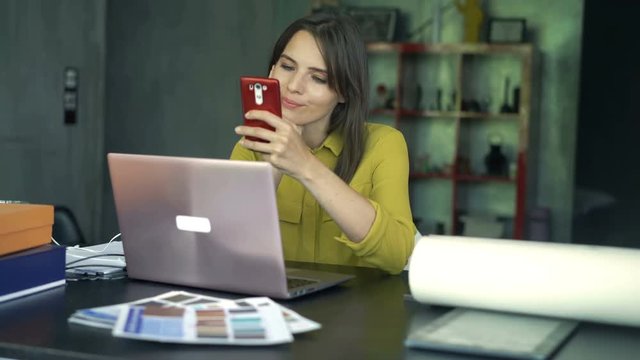 Young happy woman texting on smartphone sitting by table at home office
