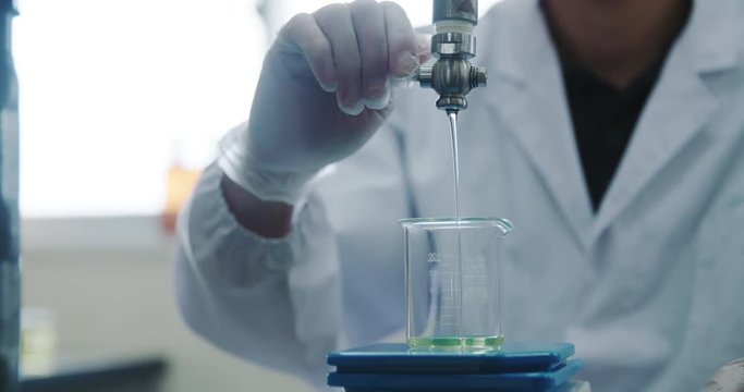 A lab technician opens a spout to pour oil into a glass beaker in a scientific laboratory