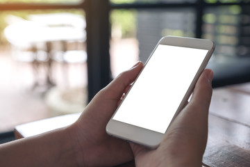 Mockup image of hands holding white mobile phone with blank screen on wooden table in cafe