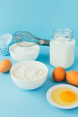 Eggs of chicken, flour in a bowl, sugar, milk in a glass jar whisk ingredient for cooking a bake on a blue background with copy space