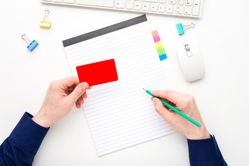 white desktop, male hands, guy writes in a notebook, holding a business card, pensil, notebook, white background with copy space, for advertisement, top view
