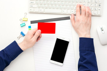 Fototapeta na wymiar white desk, male hands, guy working at the computer, holds in hand a business card, white smart phone, office supplies, Notepad, white background with copy space, for advertisement, top view