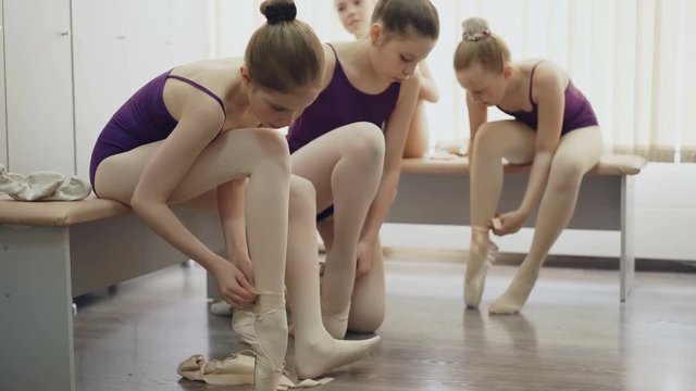 Slim girls ballet students are putting on ballet-shoes and talking sitting on benches in light locker room. Footwear, children and communication concept.