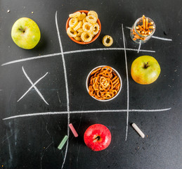 Tic tac toe healthy and unhealthy snack concept with crackers, chips and apples on black chalkboard