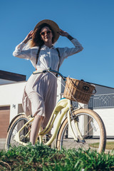 Fototapeta na wymiar Pretty girl in hat and dress by cruiser bicycle in suburb