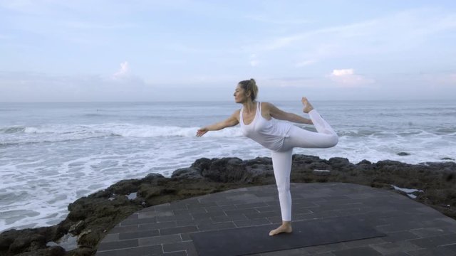 Wide shot of mature woman in white doing dancers yoga pose on coastline in morning