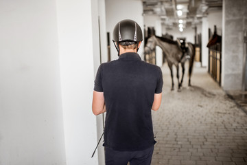 Man prepairing for horse riding standing back in the stable with horse on the backgorund