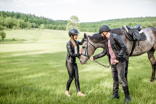 Young couple riders standing near the beautiful horse on the green meadow