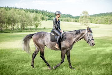 Woman in leather jacket with protective helmet riding a horse on the green meadow