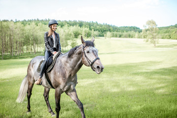 Woman in leather jacket with protective helmet riding a horse on the green meadow