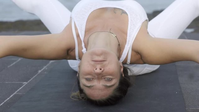 Medium shot of mature woman in white practicing yoga on beach: she lying in fish pose and stretching her hands and legs