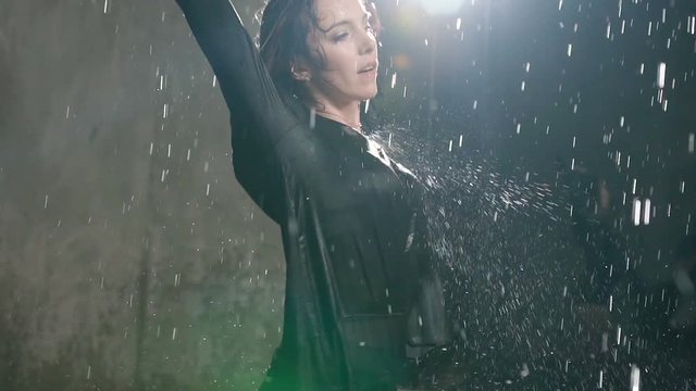 A young Caucasian girl performs a modern dance barefoot in the water under the rain drops at studio. Emotional dance. Wet ballerina dancing at the water rain in indoors with the studio light