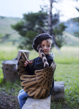 Little boy playing dress-up and camping wearing eagle wings and a raccoon hat sitting on a log by a campfire