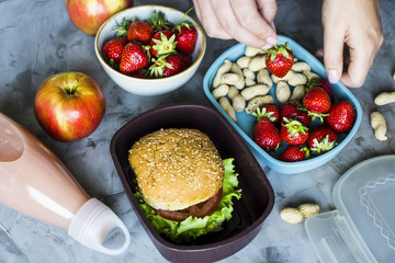 Cooking lunch for the child to school. On the gray kitchen table. Sandwich, strawberries and peanuts in lunchboxes. Top view. Woman puts strawberries in a container