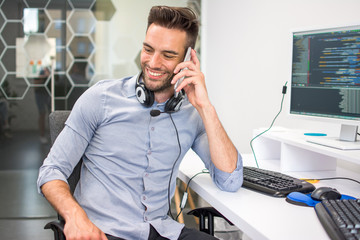 Handsome programmer with headset talking on mobile phone at his workplace.