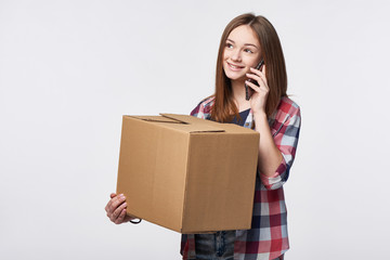 Delivery, relocation and unpacking. Smiling young woman holding cardboard box and talking on cellphone looking away at blank copy space. Call center and customer support service concept.