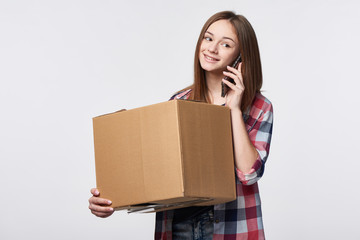 Delivery, relocation and unpacking. Smiling young woman holding cardboard box and talking on cellphone. Call center and customer support service concept.