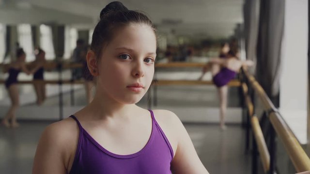 Close-up portrait of dark-haired little girl in bodysuit standing in ballet class and looking at camera. Other students are practising in background.