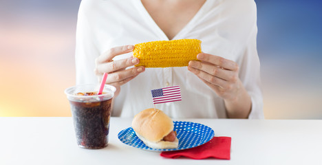 american independence day, celebration, patriotism and holidays concept - close up of woman eating corn with hot dog and cola drink at 4th july party over evening sky background