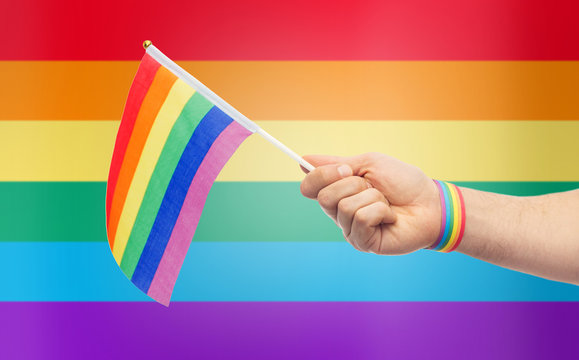 lgbt, same-sex relationships and homosexual concept - close up of male hand wearing gay pride awareness wristband holding rainbow flag