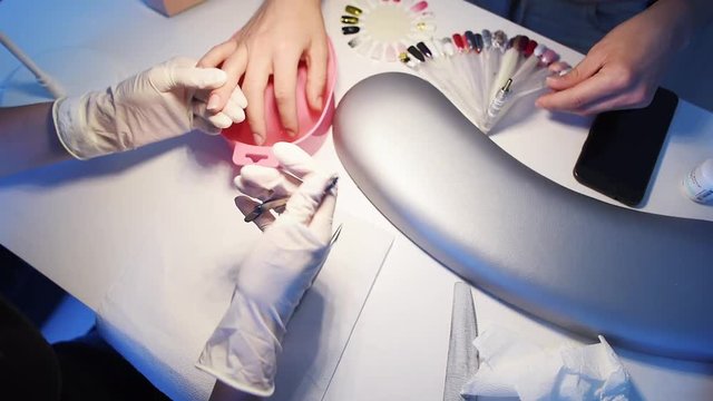 Close-up of nail master hands in disposable gloves cutting cuticles by using manicure cutters on client’s fingers while customer is choosing gel polish color from sample palette.