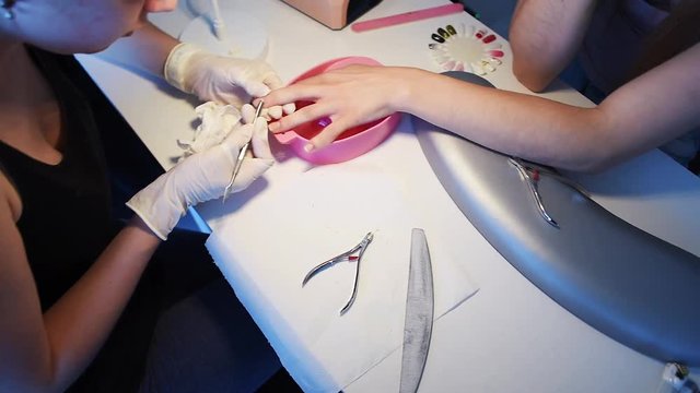 Manicurist in disposable gloves using cuticle pusher for pushing cuticles on client’s little finger. Sample palette with gel polish colors and instruments. Slow motion.