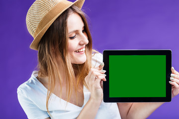 Smiling blonde girl dressed in white t-shirt and hat holds tablet computer with green screen before blue background, advertise space concept