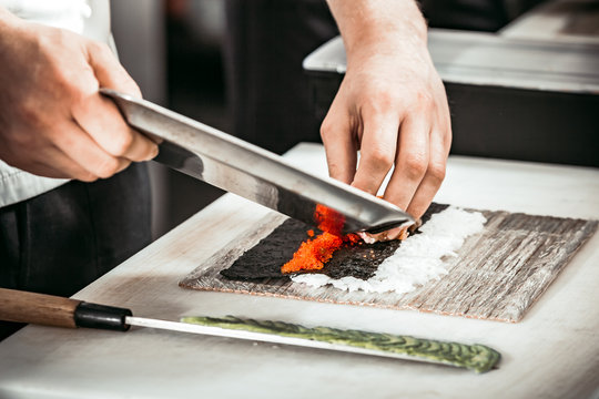 Close-up view of the man`s hands cutting the caviar on the seaweed and rice layer. The professionals are making rolls for the tasty sushi in the modern restaurant.