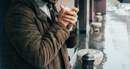 handsome man standing on the street in winter time and warming his hands. cold day. it's raining. man in jacket. Town architecture on the background. Outdoors