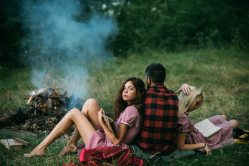 Brunette girl leaning on her boyfriend while reading book. Turn back guy looking at campfire. Young couple in love. Friends camping in woods