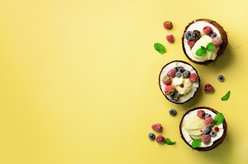 Coconut ice cream with fresh berries in coconuts halves on yellow background with copy space. Top View. Pop art design, creative summer concept. Food in minimal flat lay style.