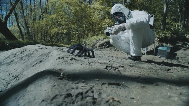 The scientist in a chemical protective suit takes pictures of a large spider on infected territory and makes notes in a notebook. Biohazard zone.
