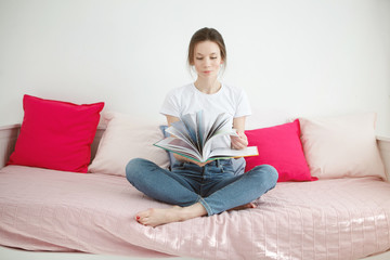 Pretty young woman enjoying reading a book at home sitting on bed smiling in pleasure in casual clothing