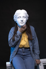 Woman wearing jeans and denim jacket posing with antique greek statue mask, fashion outfit, having fun