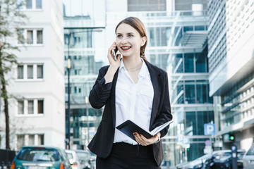 A young woman in a business suit crosses the street. Business woman talking through a mobile phone. A girl with a notepad and a phone near the office.