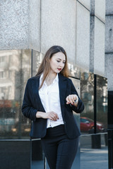 Fototapeta premium A young woman in a business suit goes to the office. A business woman checks an hour on her handwritten watch. Business Girl with Loose Hair. Business district of the city