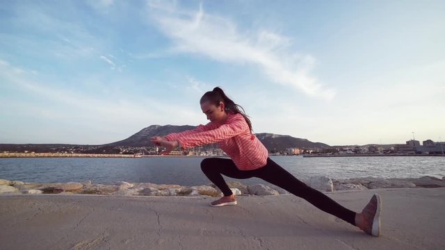 Young fit woman doing stretching exercises for legs after running by the sea with mountain on background. Slow motion.