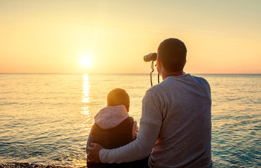 Father and son sitting on the beach and look in binocular under