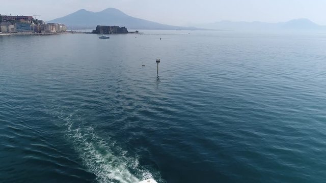 Aerial Naples flight over water cabin cruiser recreational boat camera moving towards Castel dell Ovo seaside castle located on former island of Megaride also showing Mount Vesuvius in background 4k