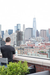Fototapeta na wymiar Man sits at the bar overlooking Lower Manhattan drinking a beer with a bottled water on the table at trendy rooftop bar in New York. Man in black shirt looking out at New York City from a rooftop bar.