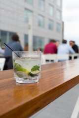 Mojito Cuban cocktail with fresh mint and black cocktail straw in a tumbler sitting on a wooden table outside on a trendy rooftop bar in New York. Table of men enjoying a drink in the background.