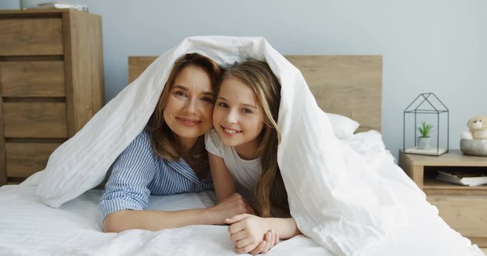 Pretty Caucasian mother and her cute daughter smiling and looking out of the blanket while lying in the bed in the morning. Upside down. Indoors