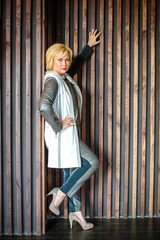 Charming blonde woman in jeans, coat and shoes stands, on brown wall background
