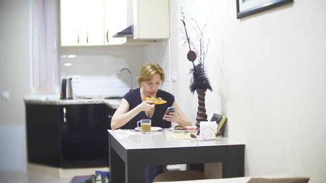 woman is eating pizza in the kitchen at home and using her smartphone, 4k, background blur