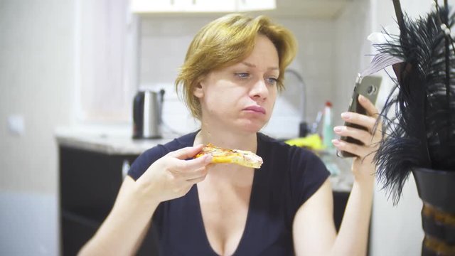woman is eating pizza in the kitchen at home and using her smartphone, 4k, background blur