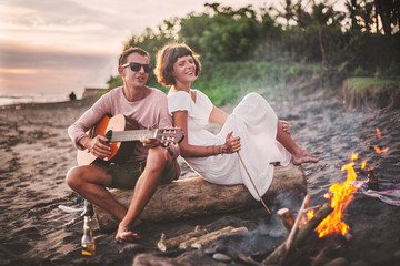 Guy with guitar and his girlfriend sitting on log and singing for his friends on summer evening by...