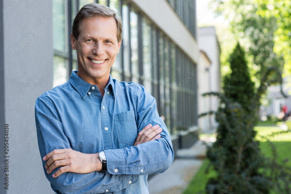 Wall mural cheerful middle aged businessman posing with crossed arms near office building